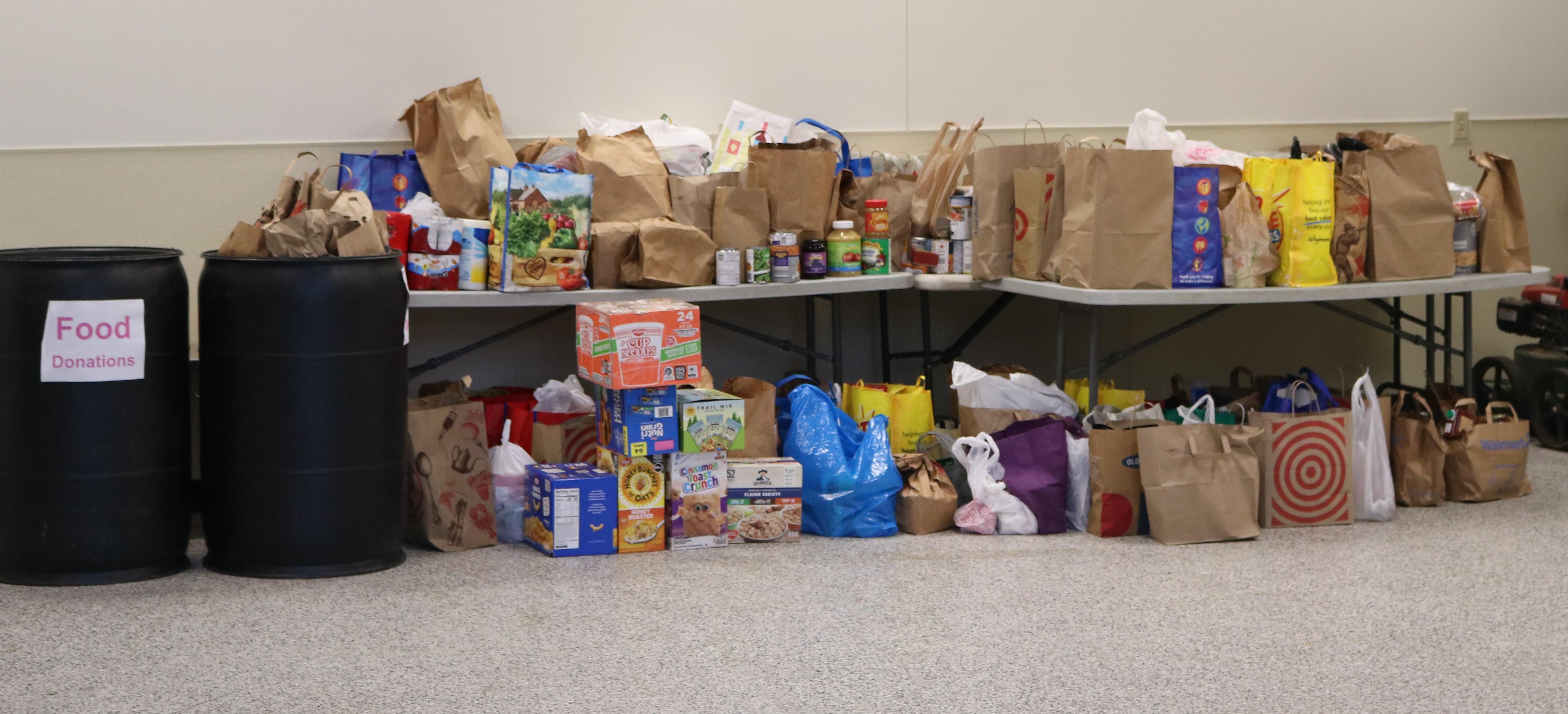 A large pile of donated non-perishable food — grocery bags, cereal boxes, and canned goods on folding tables, beside two barrels labeled Food Donations